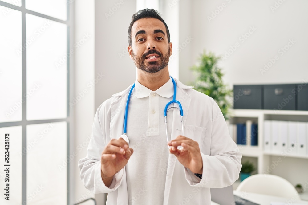 Young hispanic man wearing doctor uniform speaking at clinic