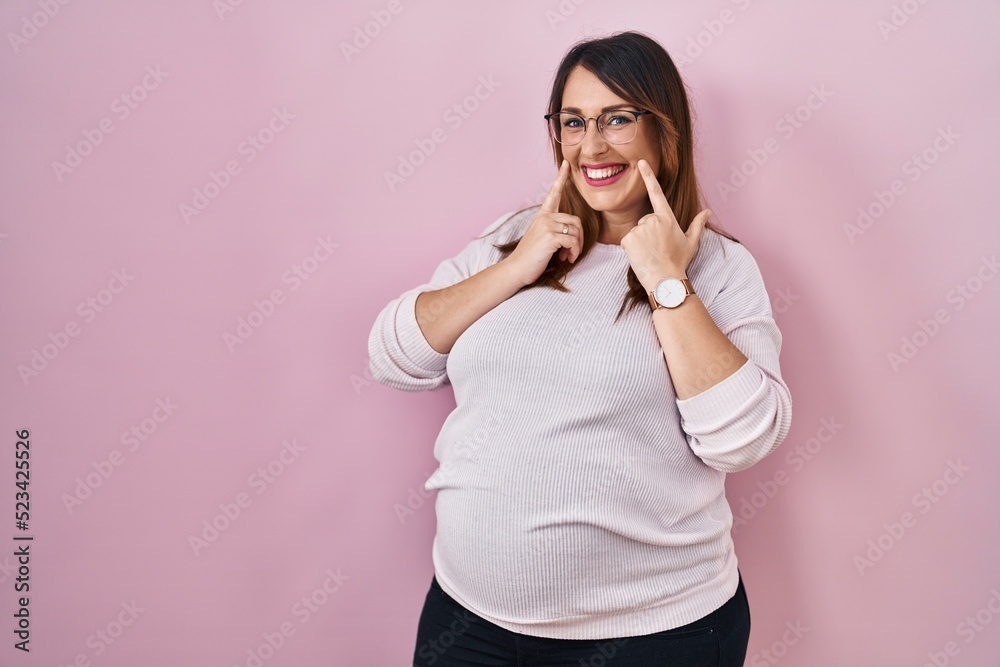 Pregnant woman standing over pink background smiling with open mouth, fingers pointing and forcing cheerful smile Pregnant woman standing over pink background smiling with open mouth, fingers pointing and forcing cheerful smile
