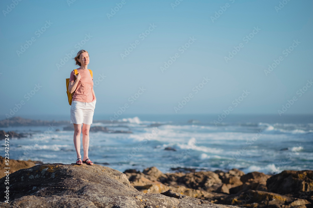 A woman with a backpack stands on the rocks near the sea.