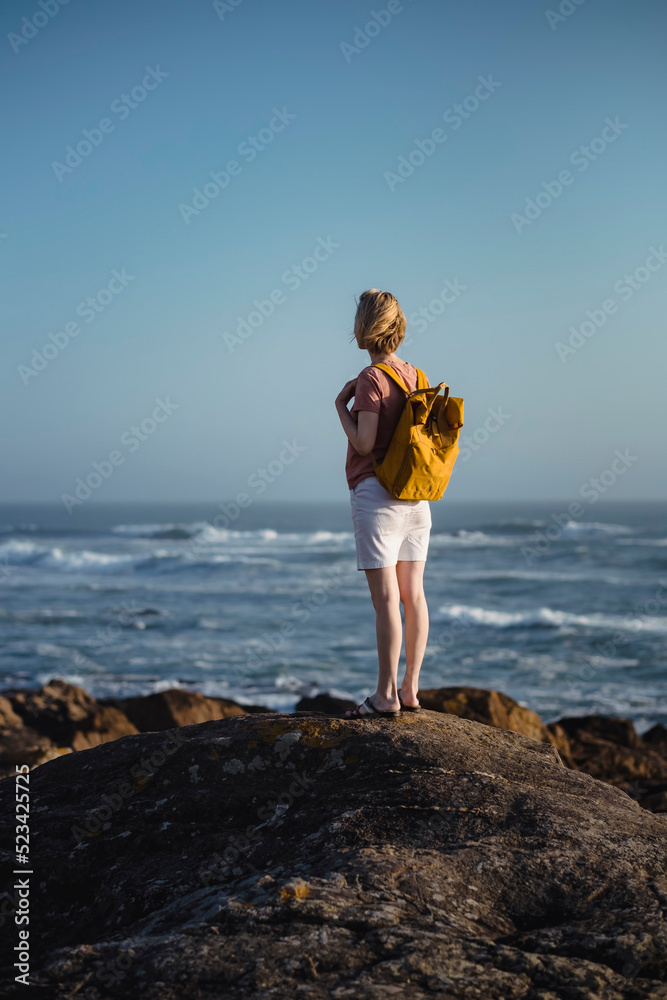 A woman with a backpack stands on the rocks near the sea. Back view.