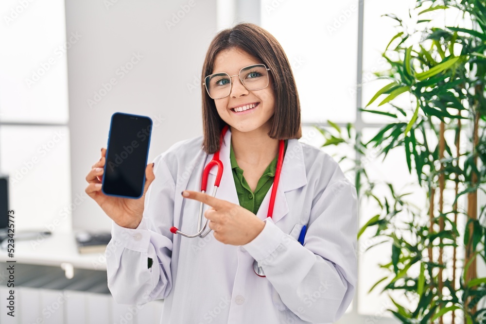 Young hispanic doctor woman showing smartphone screen smiling happy pointing with hand and finger Young hispanic doctor woman showing smartphone screen smiling happy pointing with hand and finger