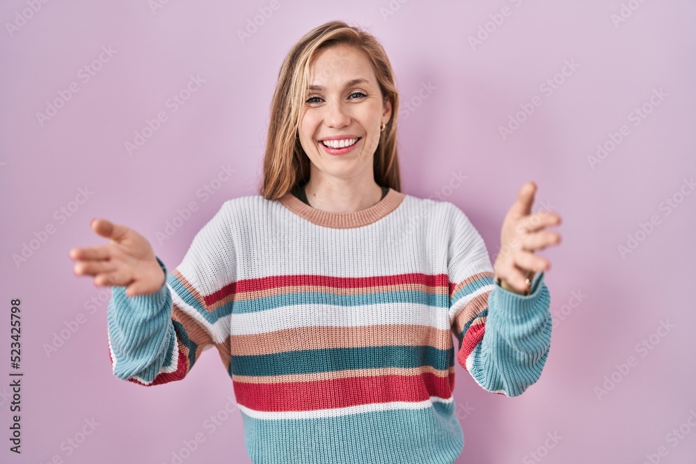 Young blonde woman standing over pink background smiling cheerful offering hands giving assistance and acceptance. Young blonde woman standing over pink background smiling cheerful offering hands giving assistance and acceptance.