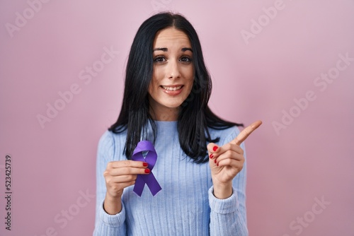 Hispanic woman holding purple ribbon awareness smiling happy pointing with hand and finger to the side