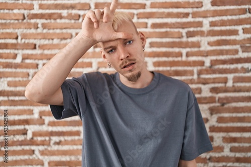 Young caucasian man standing over bricks wall making fun of people with fingers on forehead doing loser gesture mocking and insulting.