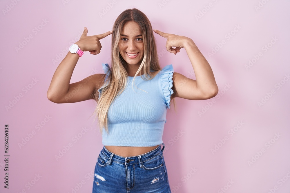 Young blonde woman standing over pink background smiling pointing to head with both hands finger, great idea or thought, good memory Young blonde woman standing over pink background smiling pointing to head with both hands finger, great idea or thought, good memory