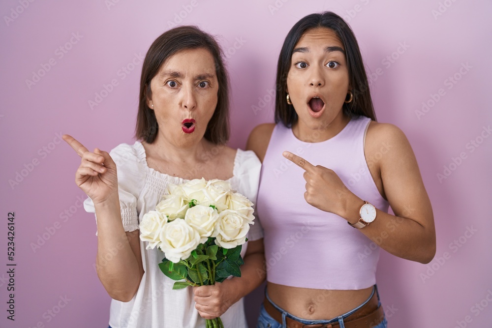 Fototapeta premium Hispanic mother and daughter holding bouquet of white flowers surprised pointing with finger to the side, open mouth amazed expression.