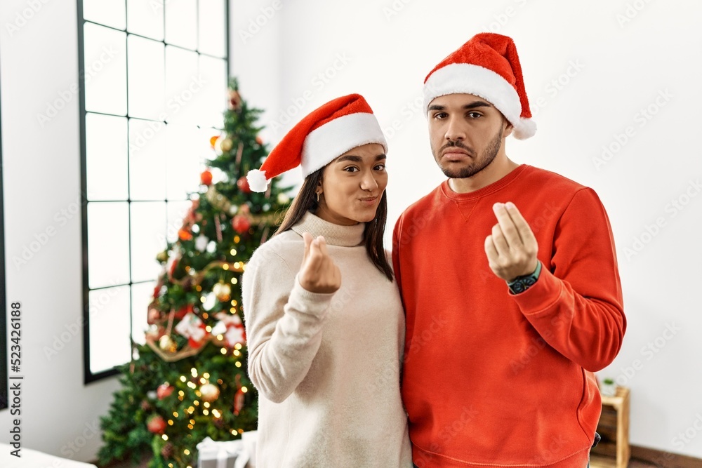 Young hispanic couple standing by christmas tree doing money gesture with hands, asking for salary payment, millionaire business Young hispanic couple standing by christmas tree doing money gesture with hands, asking for salary payment, millionaire business