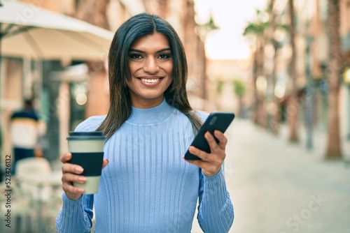 Young hispanic woman using smartphone and drinking a cup of coffee at the town