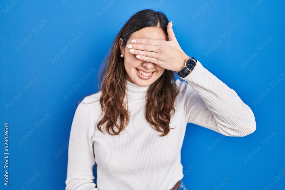 Young hispanic woman standing over blue background smiling and laughing with hand on face covering eyes for surprise. blind concept. Young hispanic woman standing over blue background smiling and laughing with hand on face covering eyes for surprise. blind concept.