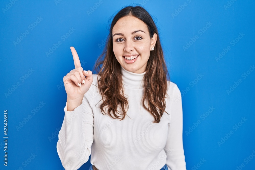 Young hispanic woman standing over blue background pointing finger up with successful idea. exited and happy. number one. Young hispanic woman standing over blue background pointing finger up with successful idea. exited and happy. number one.