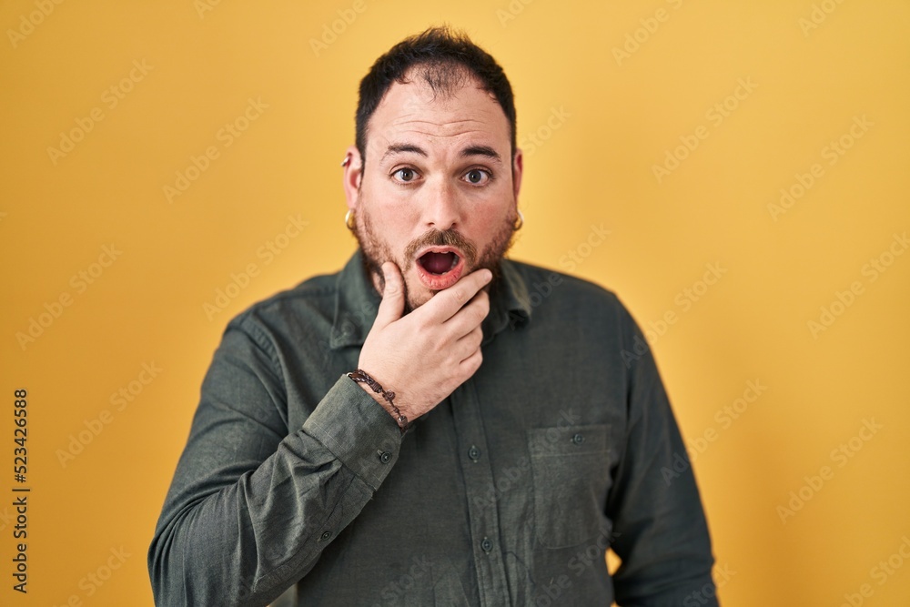 Plus size hispanic man with beard standing over yellow background looking fascinated with disbelief, surprise and amazed expression with hands on chin Plus size hispanic man with beard standing over yellow background looking fascinated with disbelief, surprise and amazed expression with hands on chin