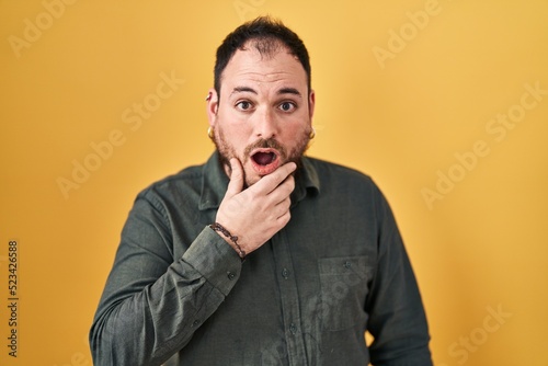 Plus size hispanic man with beard standing over yellow background looking fascinated with disbelief, surprise and amazed expression with hands on chin