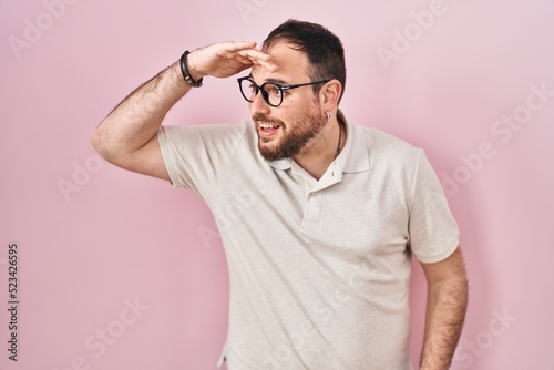 Plus size hispanic man with beard standing over pink background very happy and smiling looking far away with hand over head. searching concept.