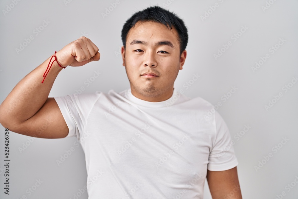 Young chinese man standing over white background strong person showing arm muscle, confident and proud of power Young chinese man standing over white background strong person showing arm muscle, confident and proud of power
