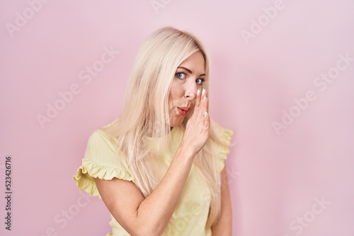 Caucasian woman standing over pink background hand on mouth telling secret rumor, whispering malicious talk conversation