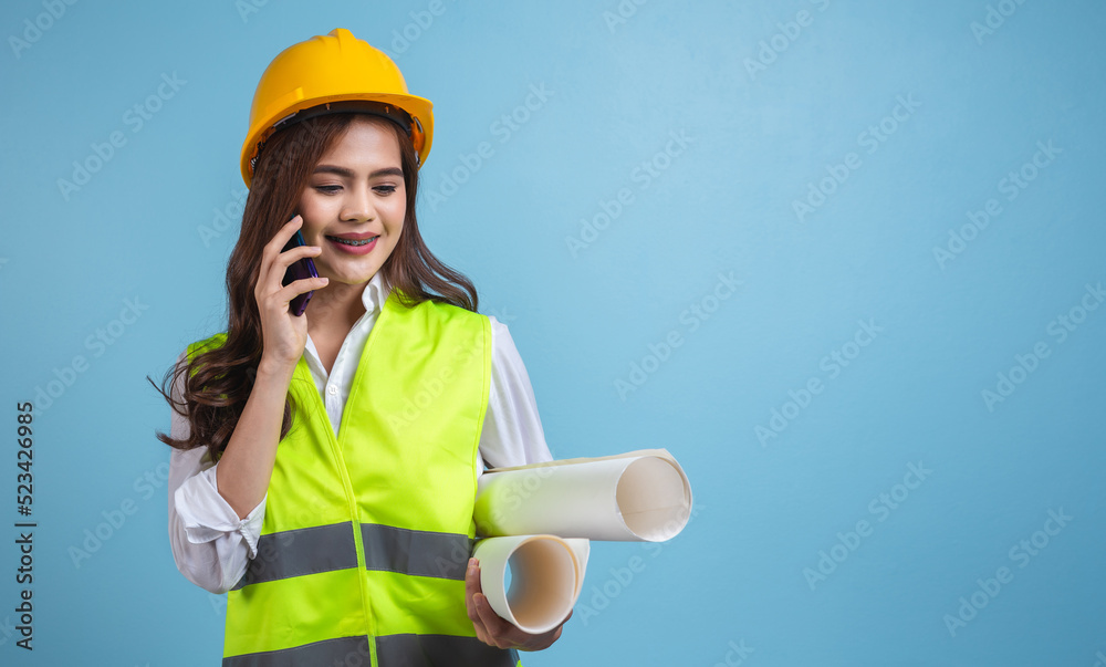 Engineer Asian woman wearing yellow safety helmet and hold blueprint using smartphone on mint blue background. Studio shot, copy space