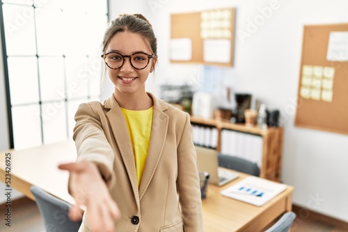 Young brunette teenager wearing business style at office smiling friendly offering handshake as greeting and welcoming. successful business.