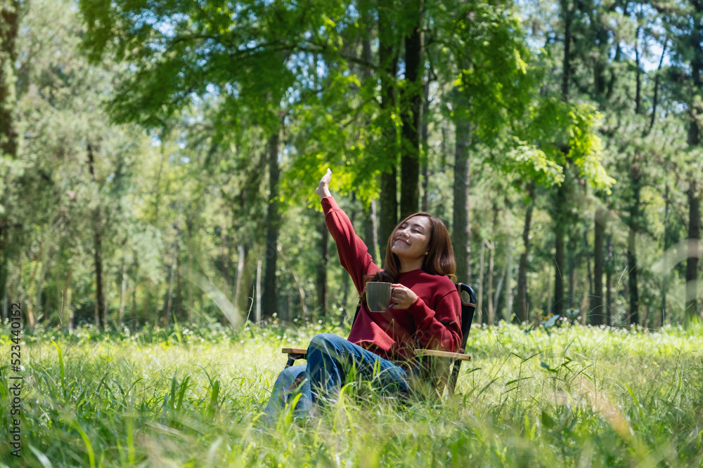 Portrait image of a happy young asian woman raising hand while drinking coffee and sitting on a chair in the park Portrait image of a happy young asian woman raising hand while drinking coffee and sitting on a chair in the park