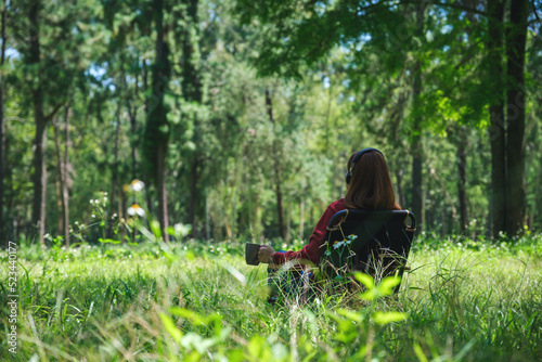 Rear view of a young woman enjoy listening to music with headphone while drinking coffee and sitting on a camping chair in the park