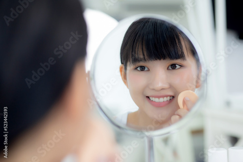 Beautiful young asian woman applying powder puff on cheek makeup of cosmetic looking mirror, beauty of girl with skin face and wrinkle in the bedroom, cosmetology for female, skincare concept.