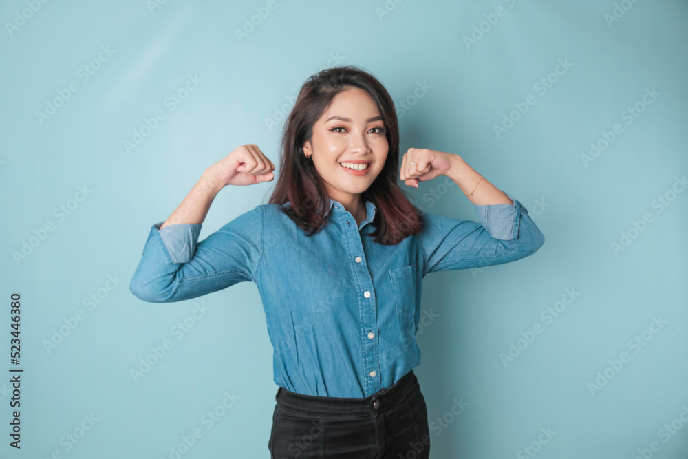 Fototapeta premium Excited Asian woman wearing a blue shirt showing strong gesture by lifting her arms and muscles smiling proudly