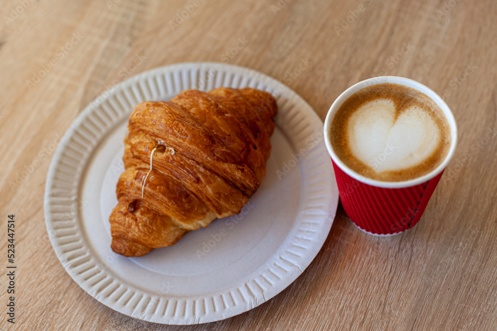 Cup of cappuccino with latte art and croissant on wooden table. Beautiful foam, red paper cup. Pastry and Coffee to go on wooden background with heart shaped latte art on top. Morning breakfast. Cup of cappuccino with latte art and croissant on wooden table. Beautiful foam, red paper cup. Pastry and Coffee to go on wooden background with heart shaped latte art on top. Morning breakfast.