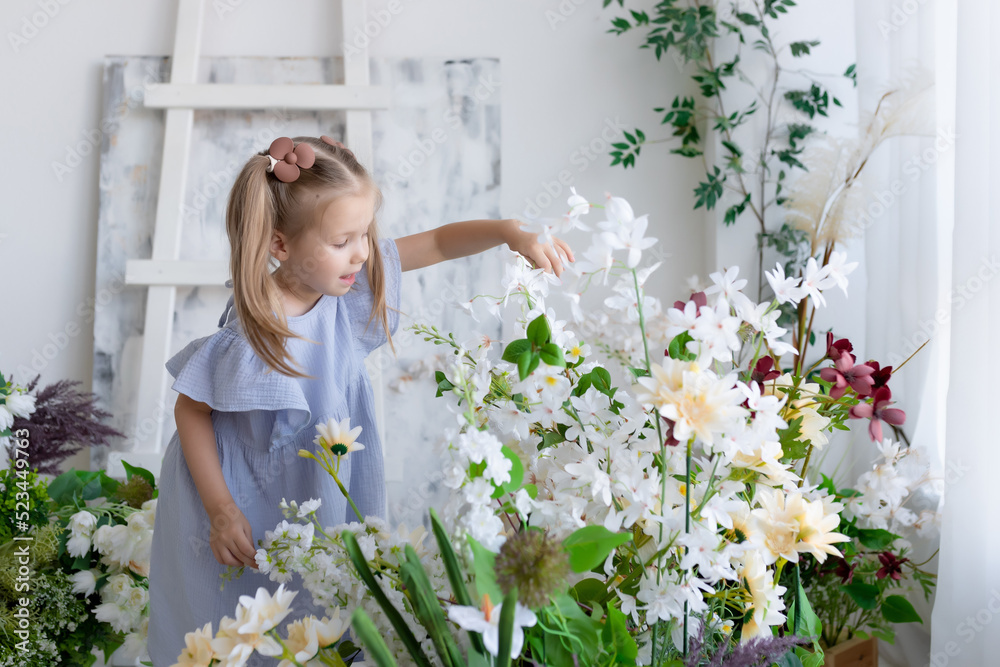 Little girl in a blue dress stands surrounded by beautiful flowers. Touching flowers with hands. Children's products Little girl in a blue dress stands surrounded by beautiful flowers. Touching flowers with hands. Children's products