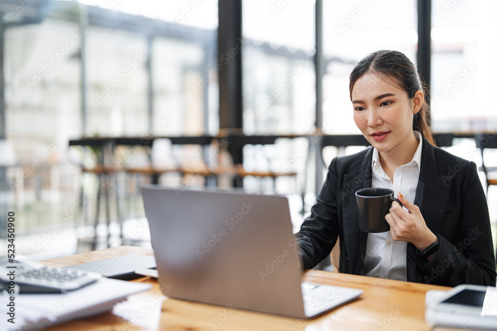 Asian businesswoman in formal suit in office holding mug working on laptop at office. Asian businesswoman in formal suit in office holding mug working on laptop at office.