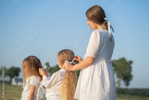 A gentle mother with two daughters in nature. Mom braids beautiful hair for her daughters. Care for beautiful and thick hair of a blonde. Psychology of the relationship between mother and daughter.