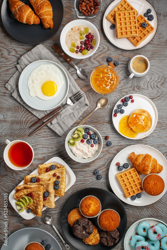 Continental breakfast captured from above (top view, flat lay). Coffee, tea, croissants, jam, egg, pancakes, maffins and oatmeal. Wooden background. Family breakfast table.