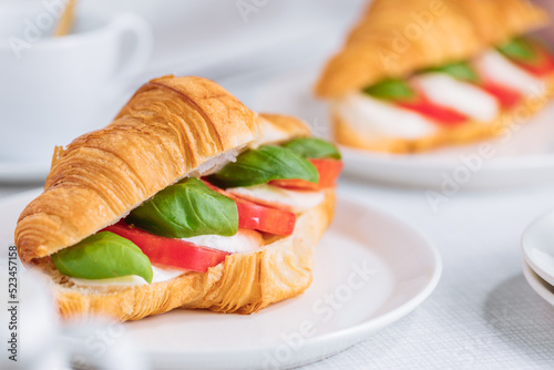 Croissant sandwich with basil, tomato and mozzarella cheese on white plate. Healthy vegetarian breakfast. Minimalistic scene, and colourful sandwich. French cuisine.