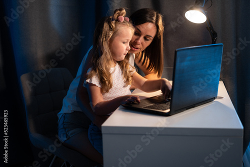Beautiful mother and child looks at laptop screen using app education program, choose cartoons in the evening, teach kid pc use, controls daughter watching on internet cyberspace