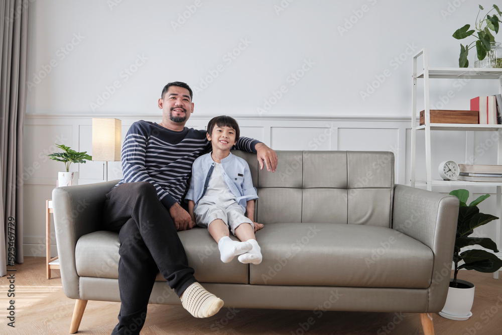 Portrait of Asian Thai family, one adult dad and little son happiness home living together, relaxing and smile, leisure on sofa in house's white room, lovely weekend and wellbeing domestic lifestyle.