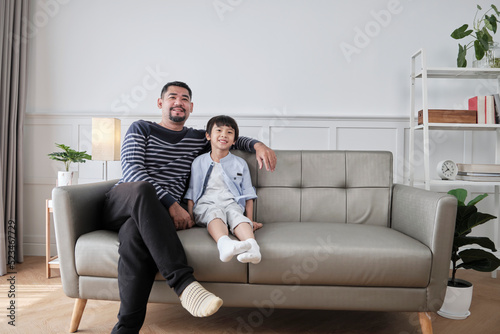 Portrait of Asian Thai family, one adult dad and little son happiness home living together, relaxing and smile, leisure on sofa in house's white room, lovely weekend and wellbeing domestic lifestyle.