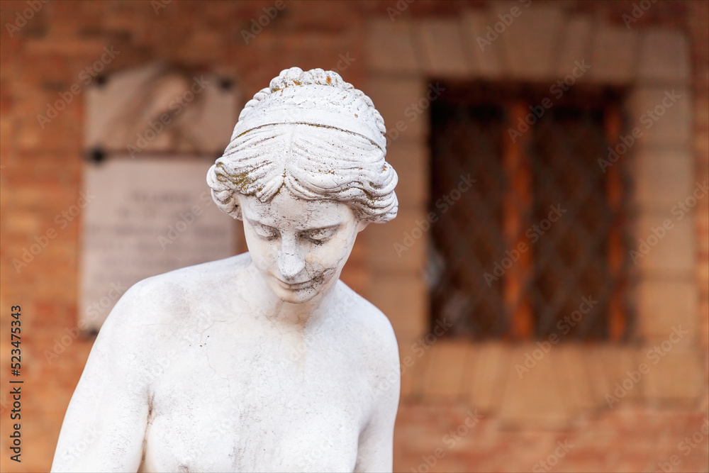 Mezzo busto di statua di donna in stile romano con sfondo una facciata in mattoncino di edificio con finestra sbarrata. 