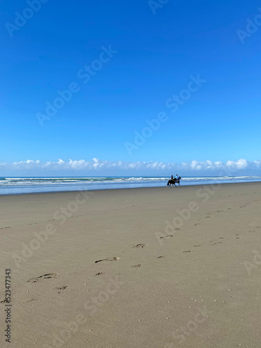 Two horseback riders on a coastline beach, Waimairi, Christchurch. Blue sky in the background. Footsteps in the sand in the foreground.