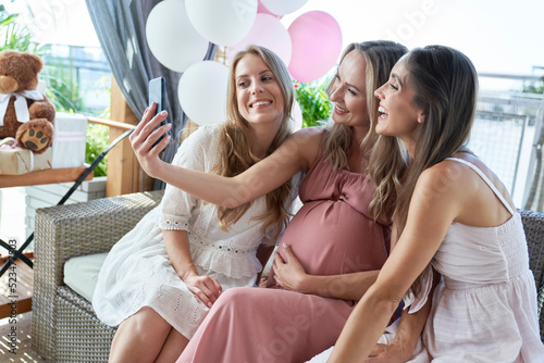 Happy pregnant woman taking selfie with friends through smart phone at baby shower