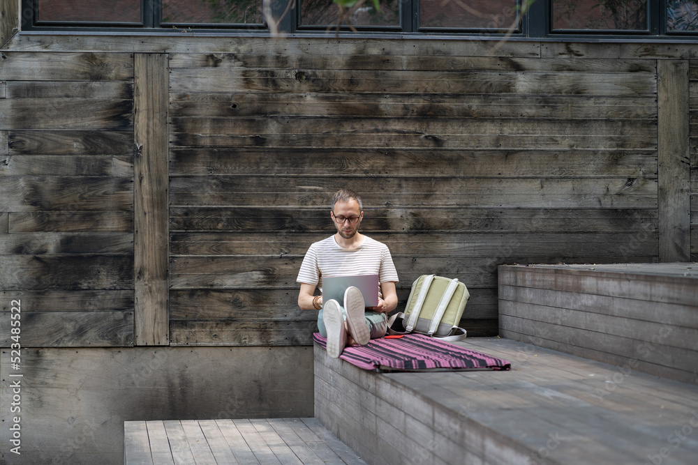 © DimaBerlin - Focused man remote worker in eyewear working on laptop remotely outside, sitting on wooden staircase at outdoor workspace. Pensive european guy student following online lectures and enjoying open air © DimaBerlin - Focused man remote worker in eyewear working on laptop remotely outside, sitting on wooden staircase at outdoor workspace. Pensive european guy student following online lectures and enjoying open air