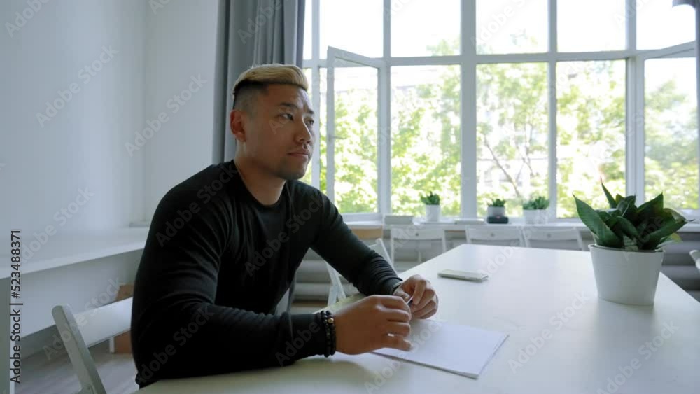 Asian blond dyed man sits at desk against large bright window listening to mentor. Young guy employee enjoys taking part in business training closeup Asian blond dyed man sits at desk against large bright window listening to mentor. Young guy employee enjoys taking part in business training closeup
