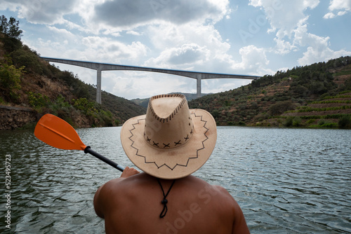 Jovem desportista de chapéu a praticar desporto com o seu caiaque no rio com uma ponte ao fundo num dia nublado photo