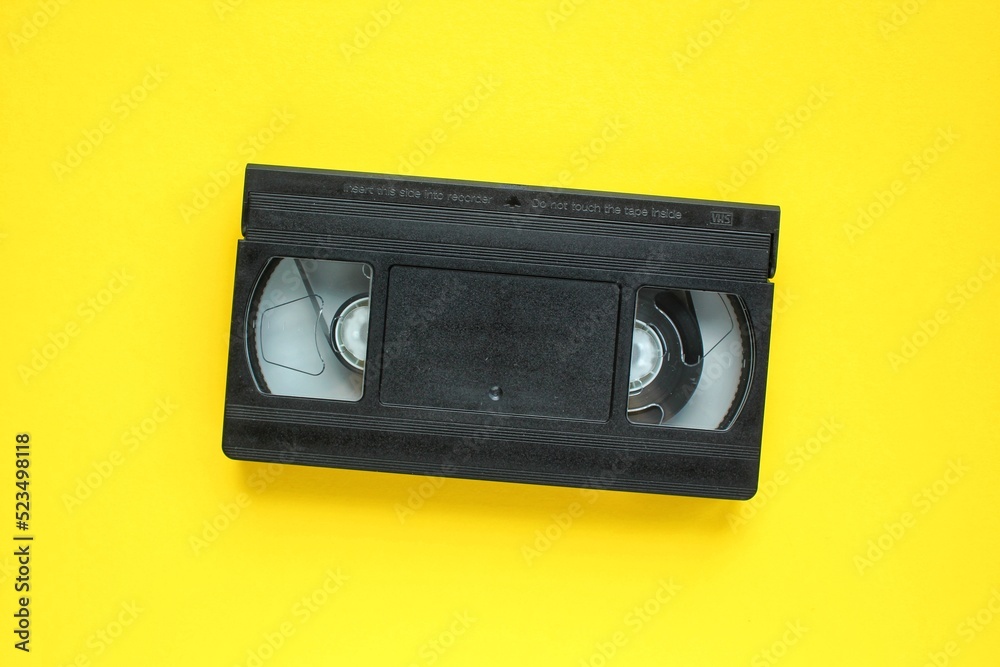 Black VHS videotape recorder cassette on yellow background. Old obsolete technology for tape recording and watching media movies. Retro, vintage, history, nostalgia concept. Top view, flat lay