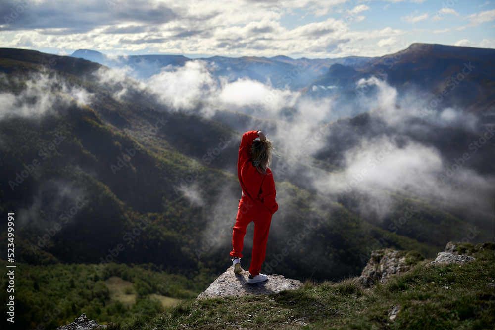 Fototapeta premium A young girl in red stands on the edge of a cliff in the mountains and looks at the clouds that have descended to the Alpine meadows