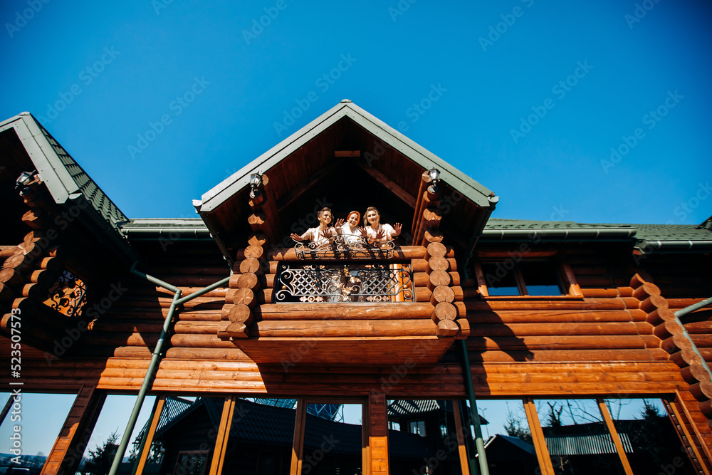 Happy red-haired bride posing with her bridesmaids on the balcony of a wooden cottage on a sunny day