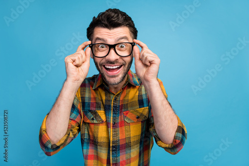 Photo of ecstatic cheerful handsome man dressed checkered colored shirt hands touching glasses staring isolated on blue color background