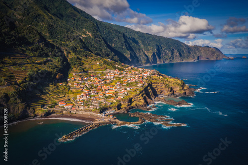 Amazing aerial view of red roofed houses located amidst green fields near Seixal beaches washing by ocean in Madeira, Portugal. October 2021