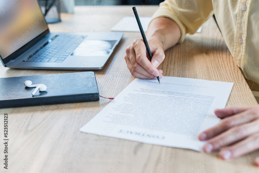 Cropped view of businessman writing on blurred contract near notebook and earphones on table. Cropped view of businessman writing on blurred contract near notebook and earphones on table.