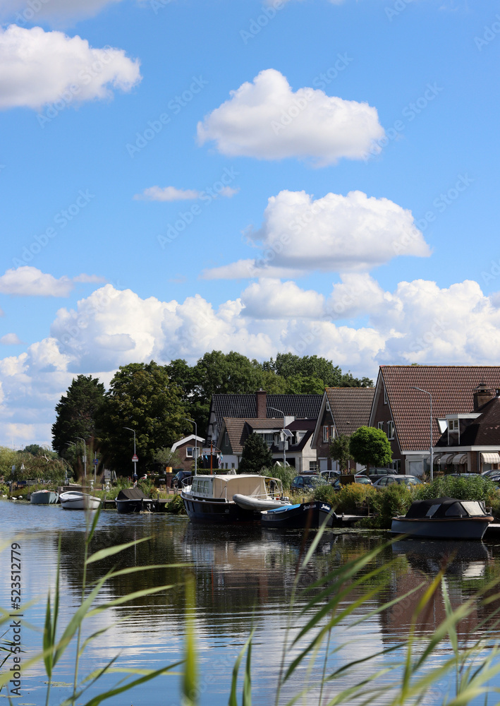 Obraz premium Dutch town on a river. Wooden houses, boats on water, green trees, cloudy sky. Country side living in the Netherlands. 