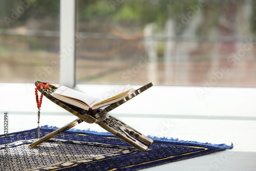 Rehal with open Quran and Misbaha on Muslim prayer rug near window indoors, space for text