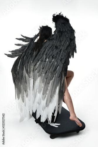 full length portrait of beautiful asian model with dark hair, wearing black gothic skirt costume, angel feather wings with horned headdress. crouching pose  isolated on studio background.
