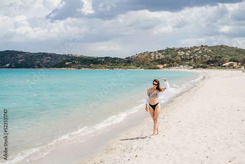A young happy woman with a beautiful figure in a swimsuit walks along the white sand beach near the blue sea. 